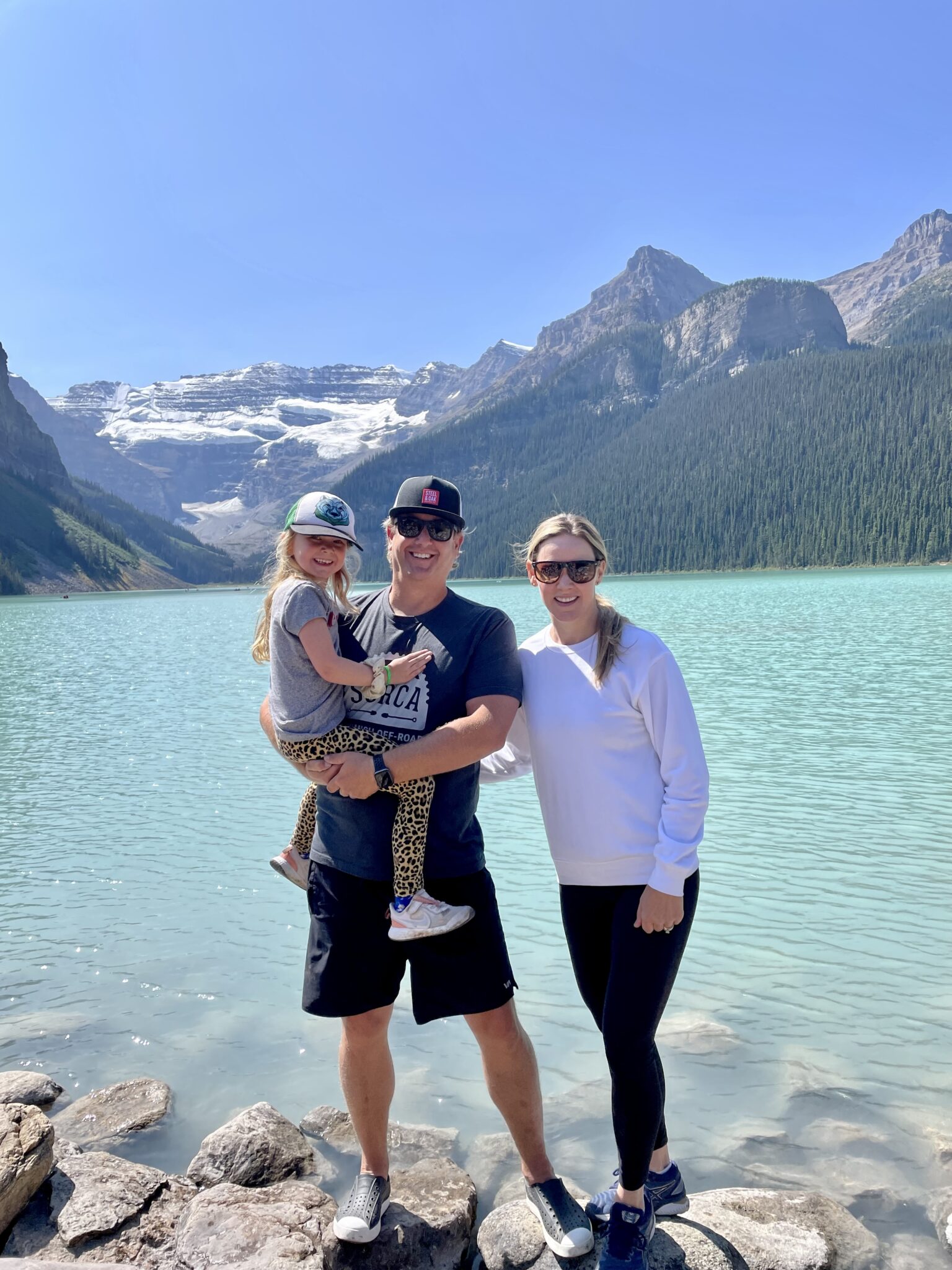A photo of Paul holding his child, with his wife standing beside him. The background is a scenic, turquoise lake and mountain in the distance.